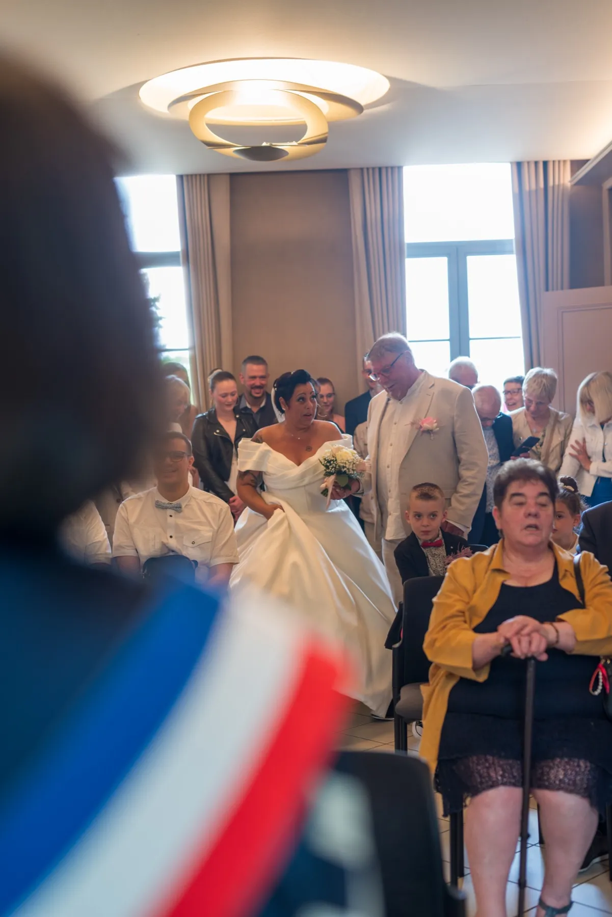 Derrière une écharpe tricolore au premier plan, la mariée remonte l’allée à côté d’un homme en costume clair