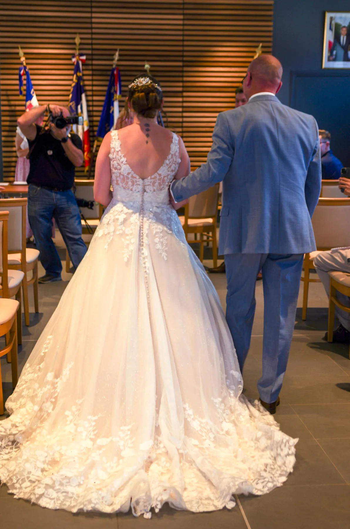 Vue de dos, la mariée avance au bras de son papa, entre les drapeaux de la mairie
