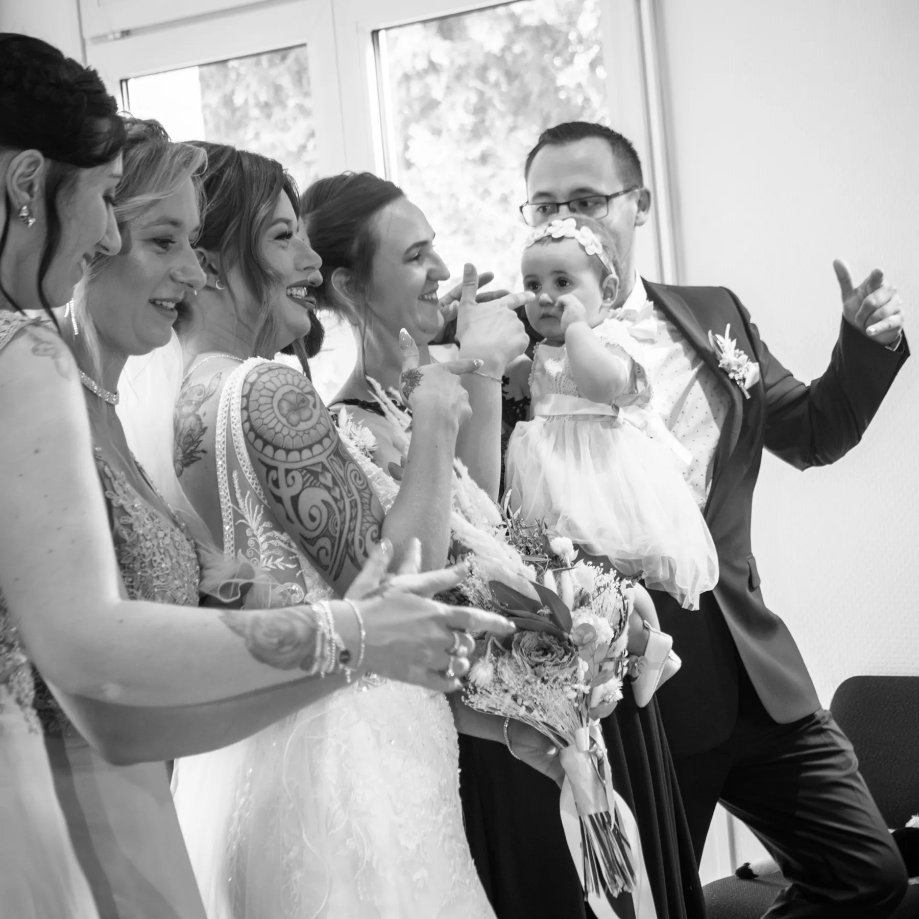 En noir et blanc, la mariée et ses proches font des signes à un bébé en robe blanche