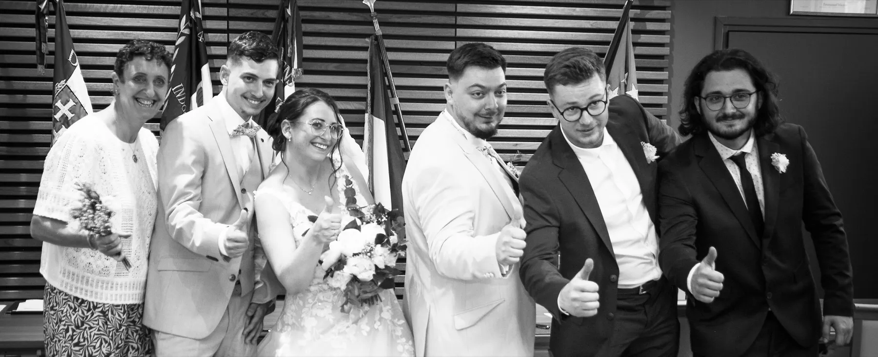 En noir et blanc, les mariés et quatre proches lèvent le pouce, devant les drapeaux