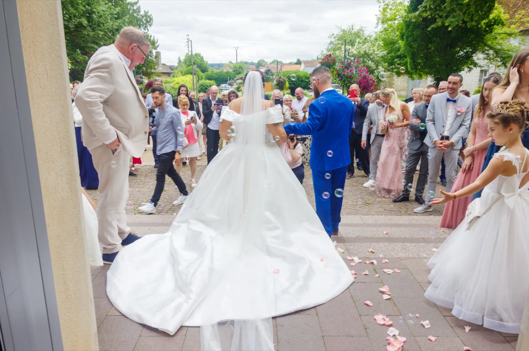 Vus de dos, les mariés s’avancent vers leurs invités, des pétales sur le pavé