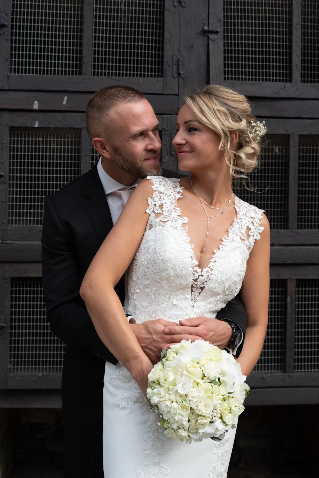 La mariée sourit tendrement, enlacée par derrière par le marié, un bouquet blanc devant eux