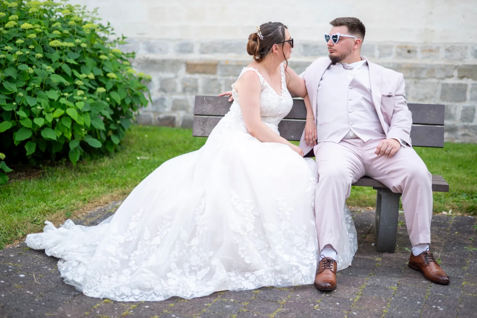 Assis sur un banc, les mariés portent des lunettes de soleil en forme de cœur et se regardent