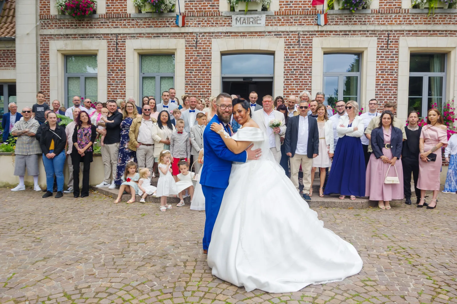 Devant la façade en briques de la mairie, les mariés s’enlacent, entourés de tous leurs invités