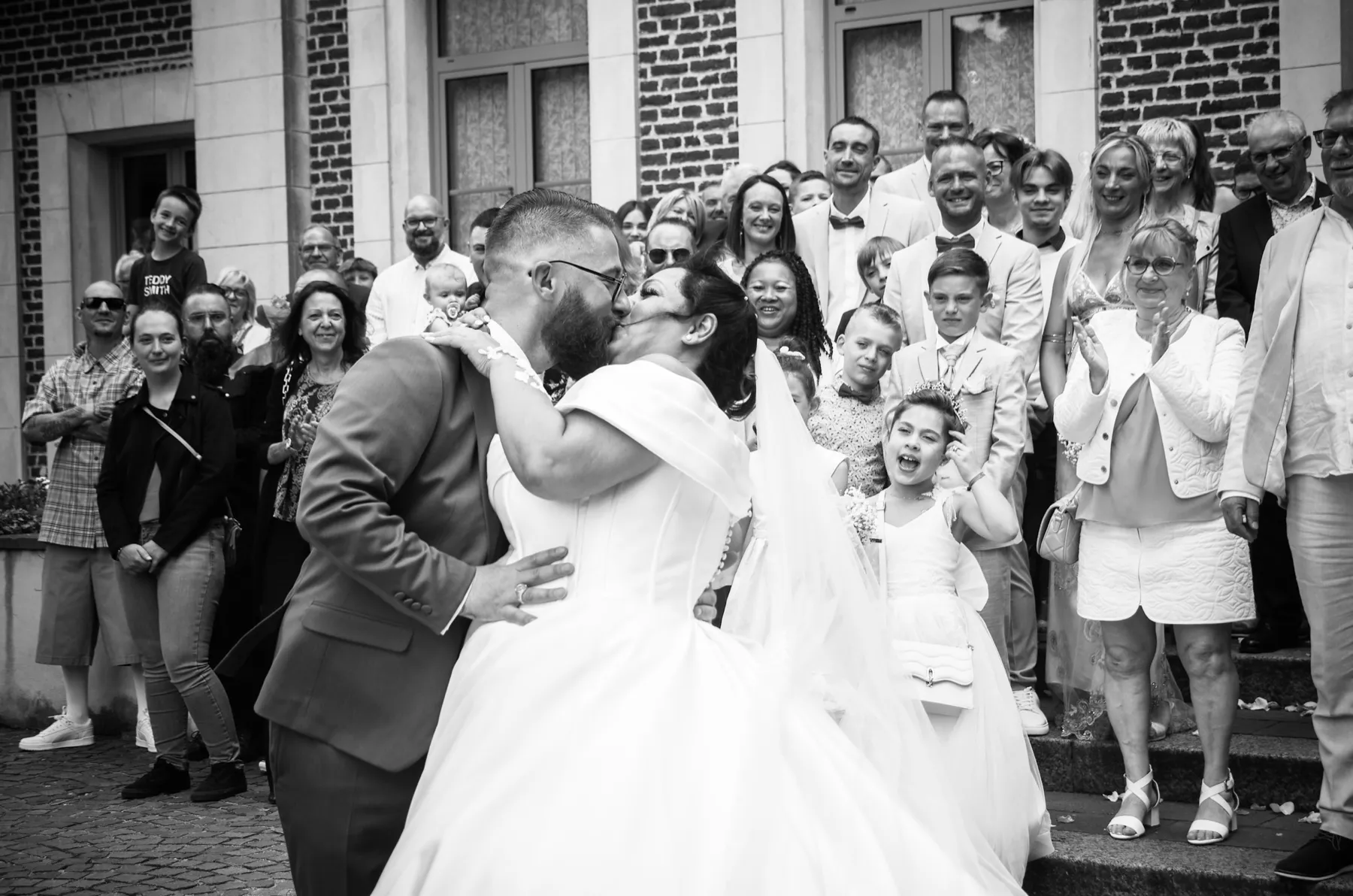En noir et blanc, les mariés s’embrassent devant leurs invités qui applaudissent