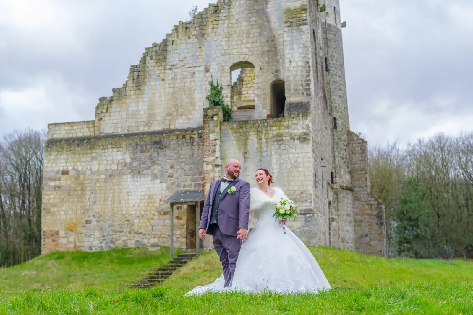 Les mariés se tiennent par la main devant des ruines en pierre, la mariée rit au vent
