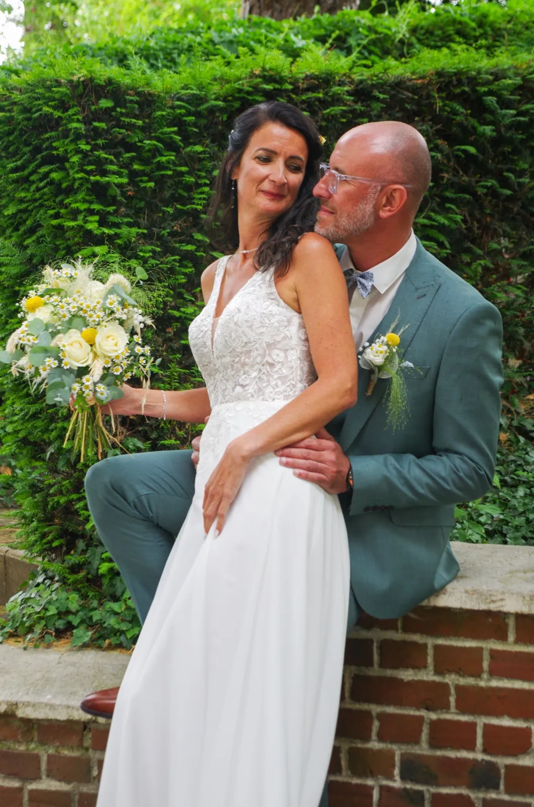 Assise sur un muret en briques, la mariée tient son bouquet jaune et blanc, le marié derrière elle