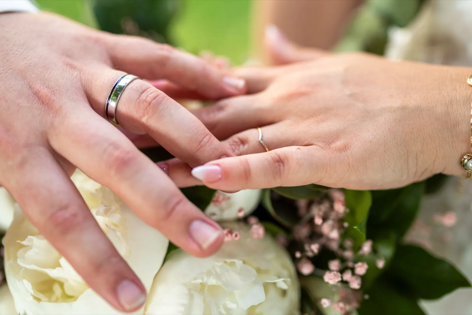 Gros plan des mains des mariés avec leurs alliances posées sur un bouquet de pivoines