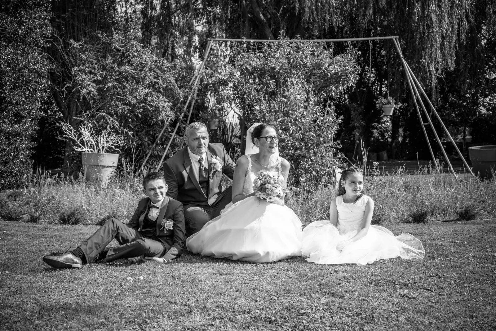 En noir et blanc, les mariés et deux enfants sont assis sur l'herbe devant une balançoire