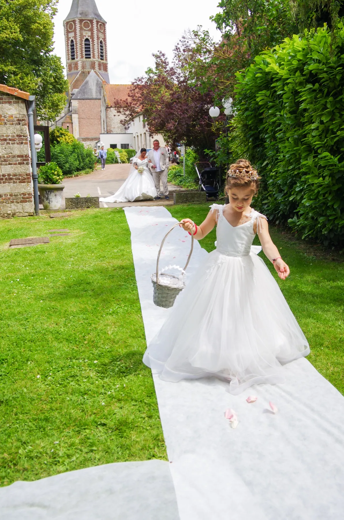 Une petite fille en robe blanche avance sur l’allée, un panier à la main, la mariée au loin