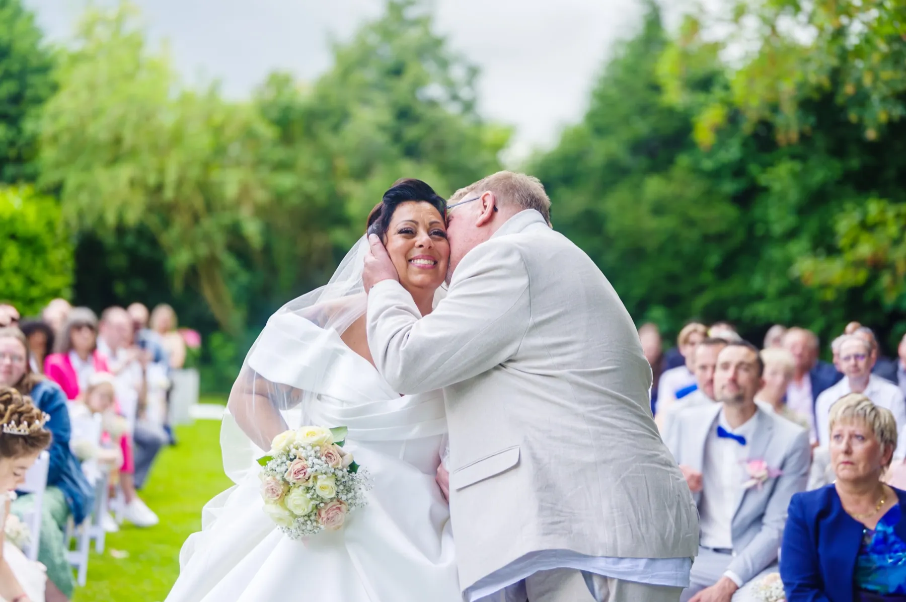 Devant les invités assis, un homme en costume clair embrasse la mariée sur la joue