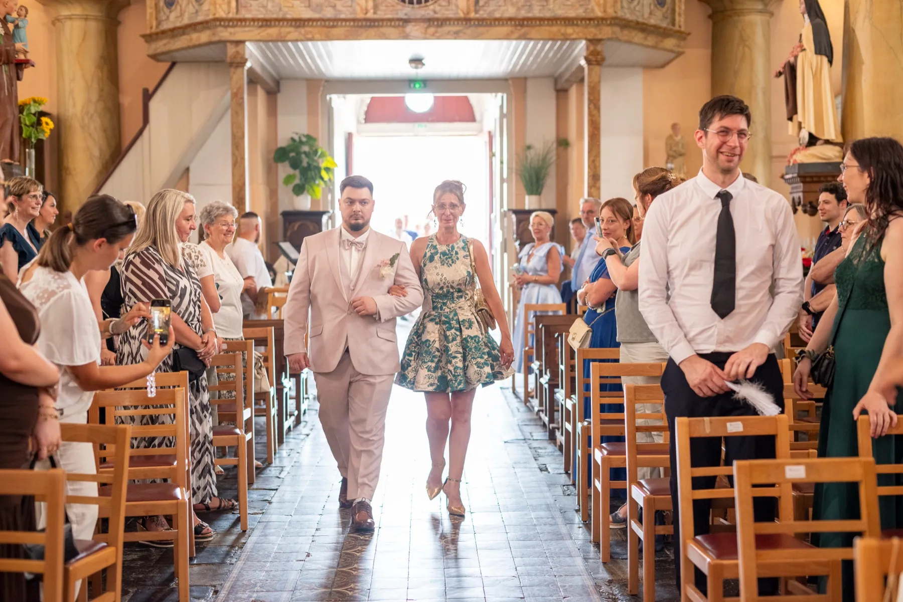 Le marié remonte l’allée de l’église au bras d’une femme en robe à fleurs