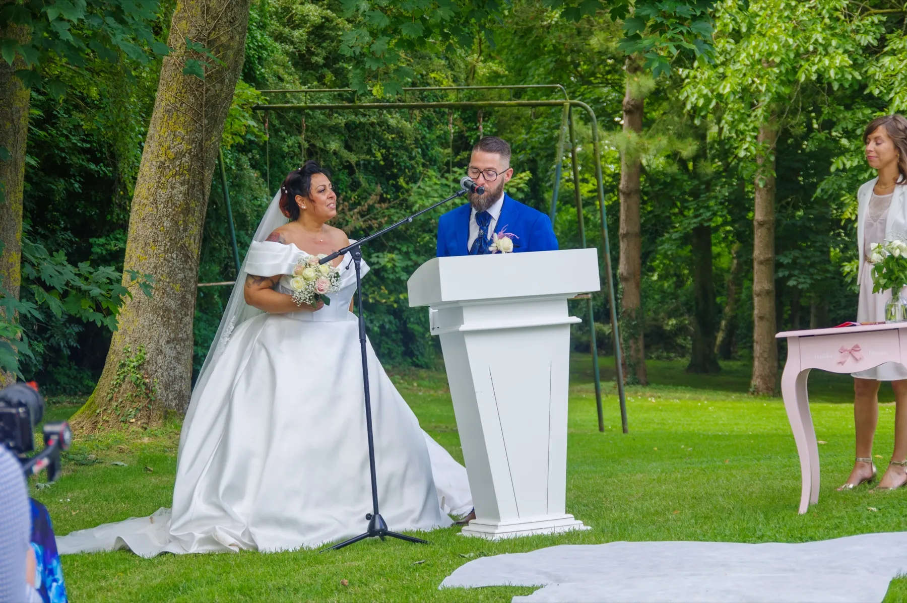 Sous les arbres, la mariée parle au micro, le marié debout derrière un pupitre blanc