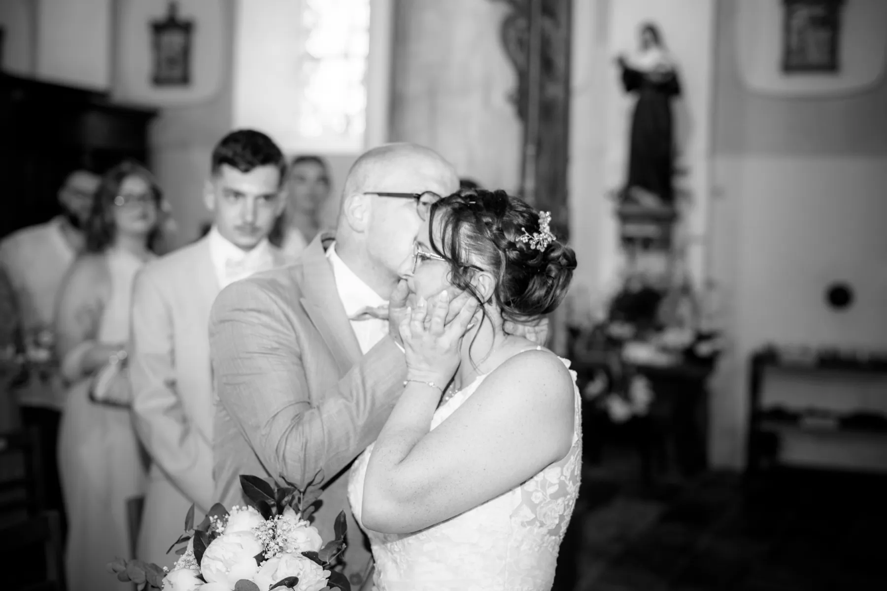 En noir et blanc, dans l’église, un homme en costume prend le visage de la mariée entre ses mains
