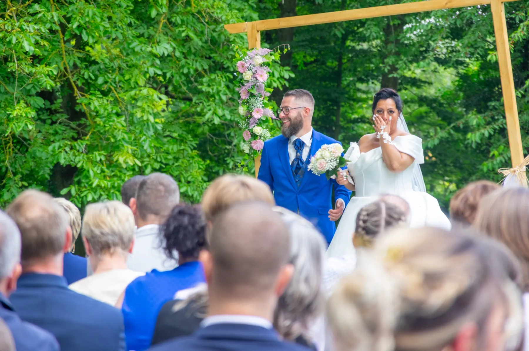 Vue par-dessus les invités, la mariée essuie une larme près du marié, sous une arche fleurie