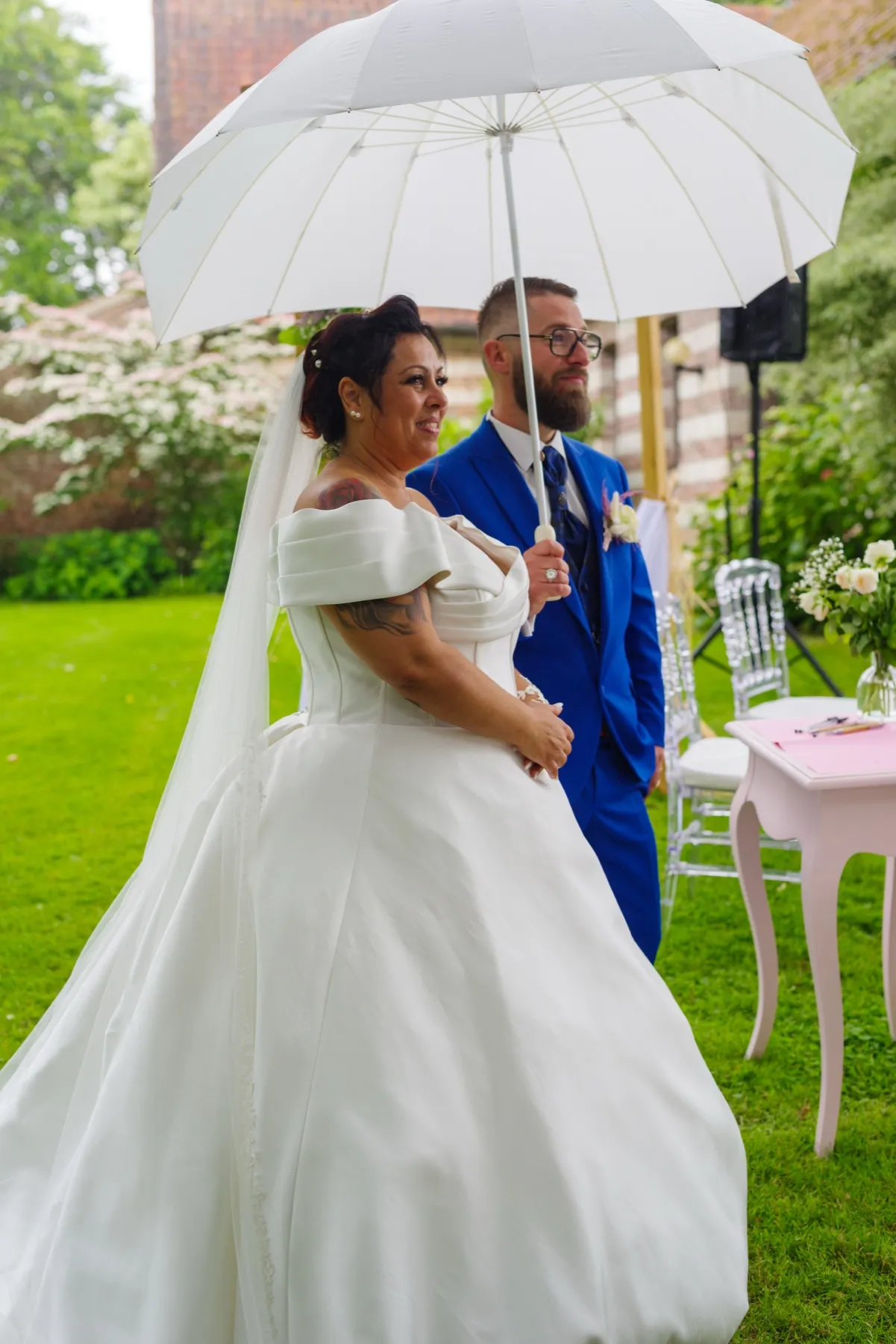La mariée dans sa grande robe et le marié sous une ombrelle blanche, dans le jardin