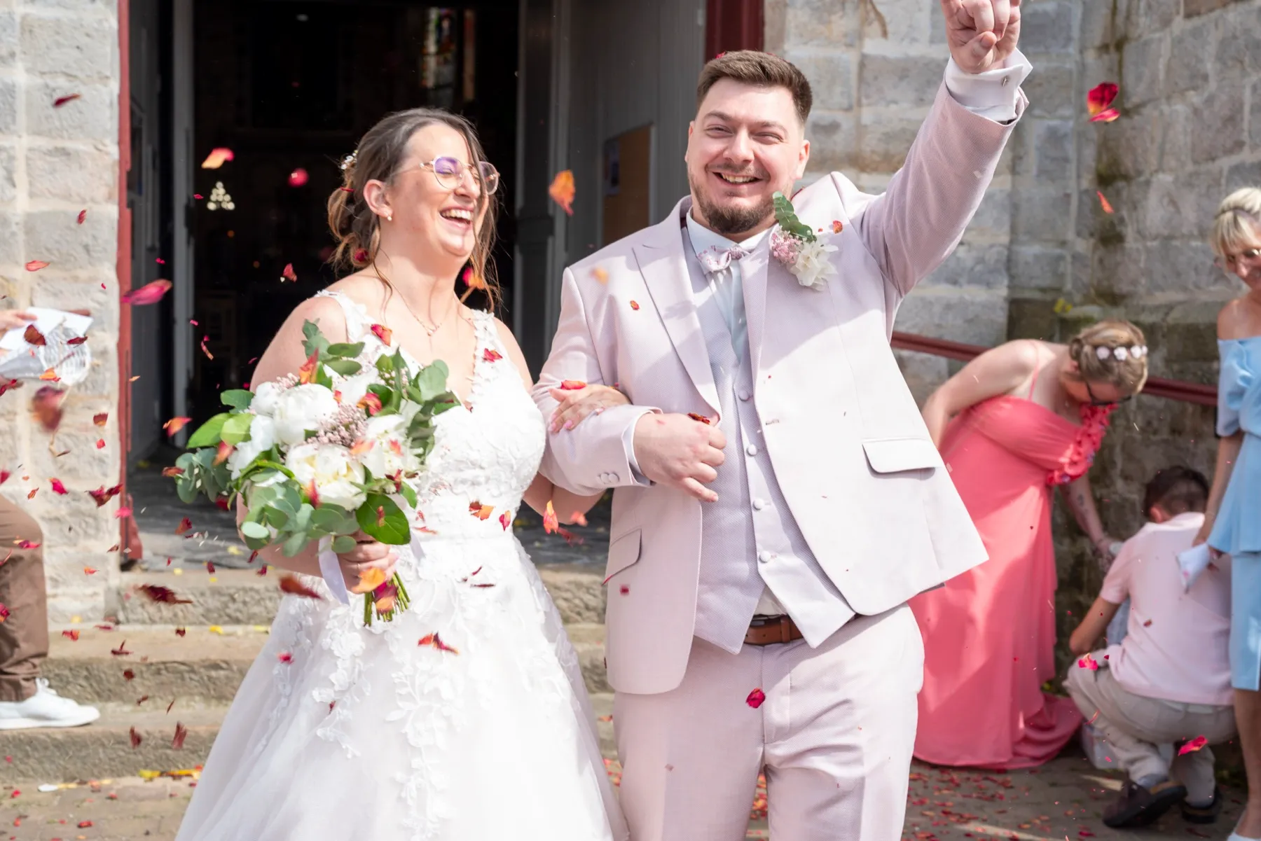 Sur le parvis, le marié lève le poing, la mariée rit sous les pétales