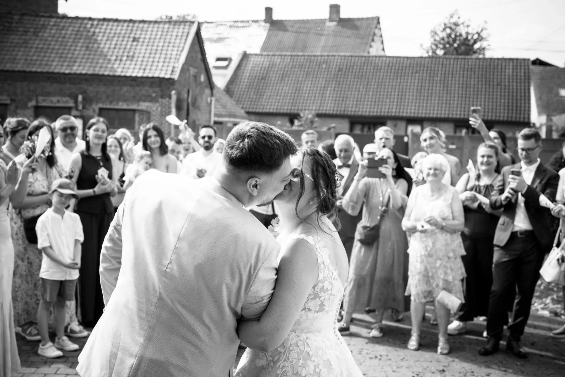 En noir et blanc, les mariés s’embrassent devant leurs invités qui les photographient