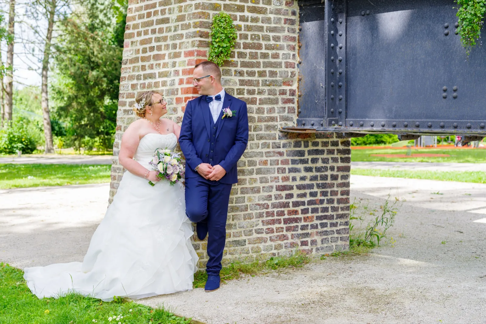 La mariée regarde le marié adossé à un pilier en briques, dans un parc ensoleillé