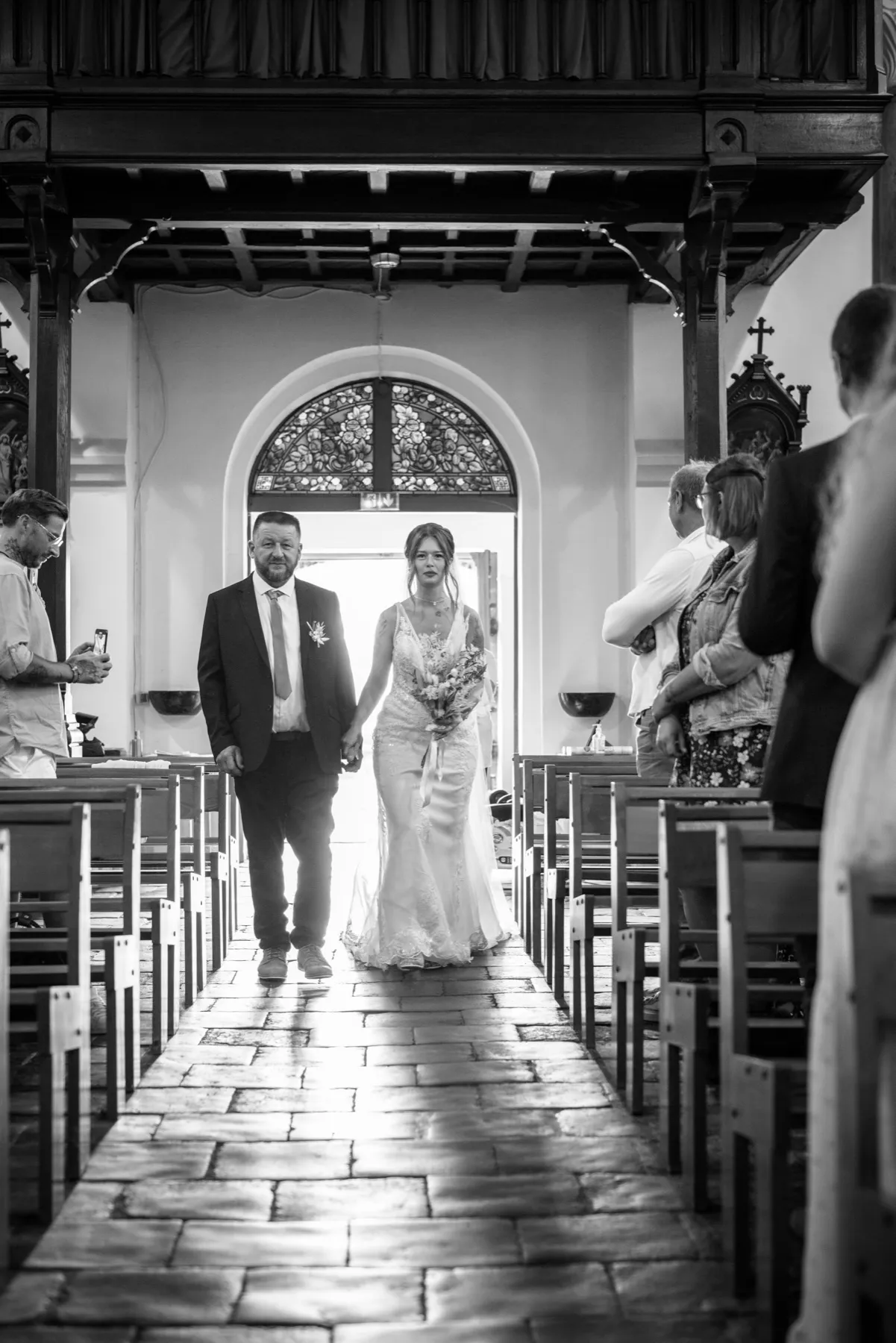 En noir et blanc, la mariée remonte l’allée de l’église en tenant la main d’un homme en costume