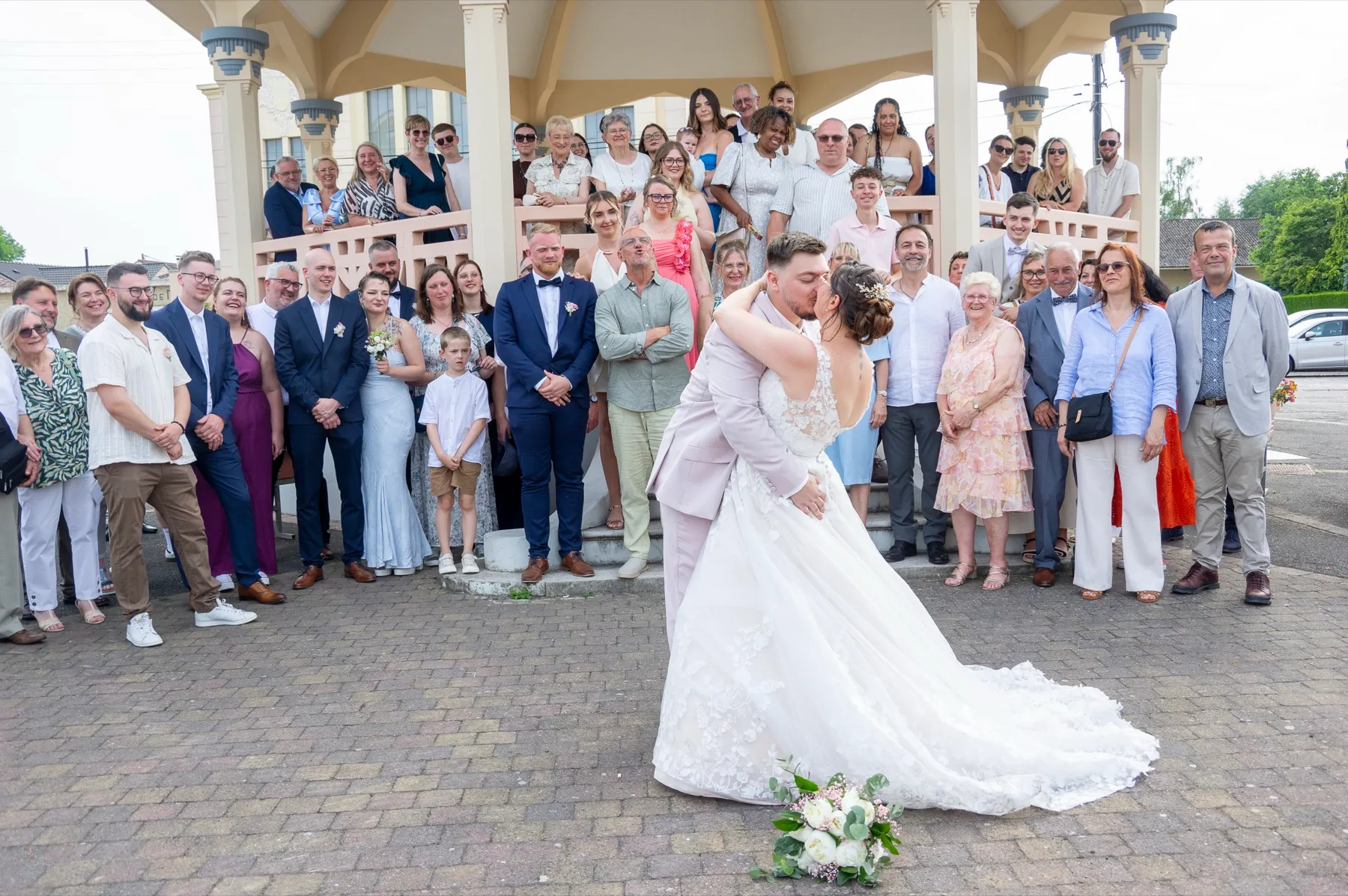 Devant le kiosque de Haillicourt, le marié renverse la mariée pour un baiser, entourés de leurs invités