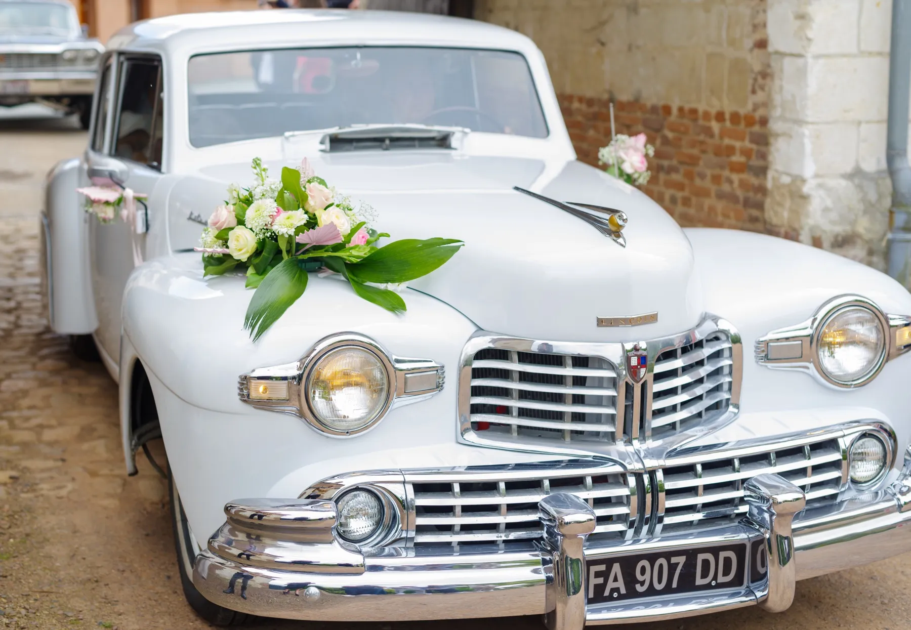 Une voiture ancienne blanche, un bouquet de fleurs sur le capot, devant un mur de briques