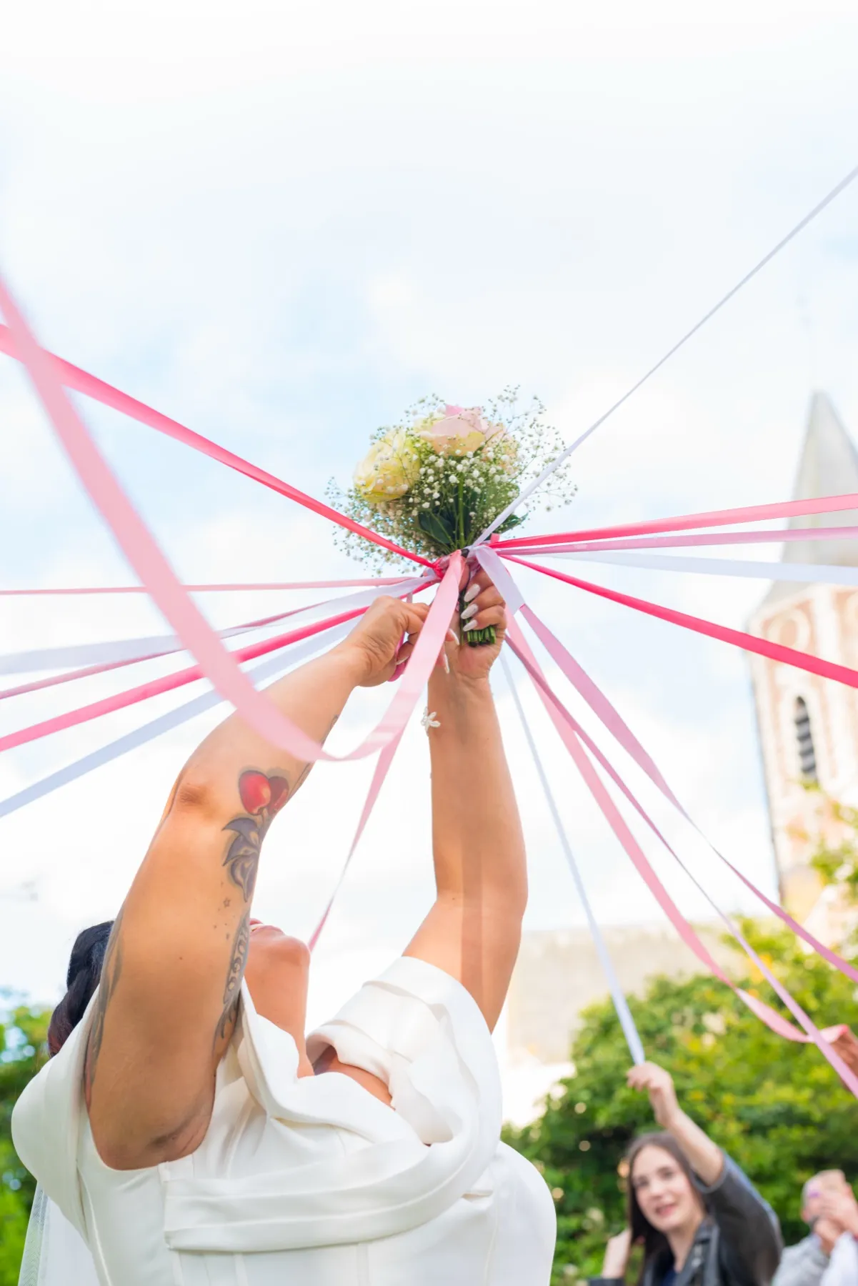 La mariée lève son bouquet, d’où partent des rubans roses et blancs vers les invités