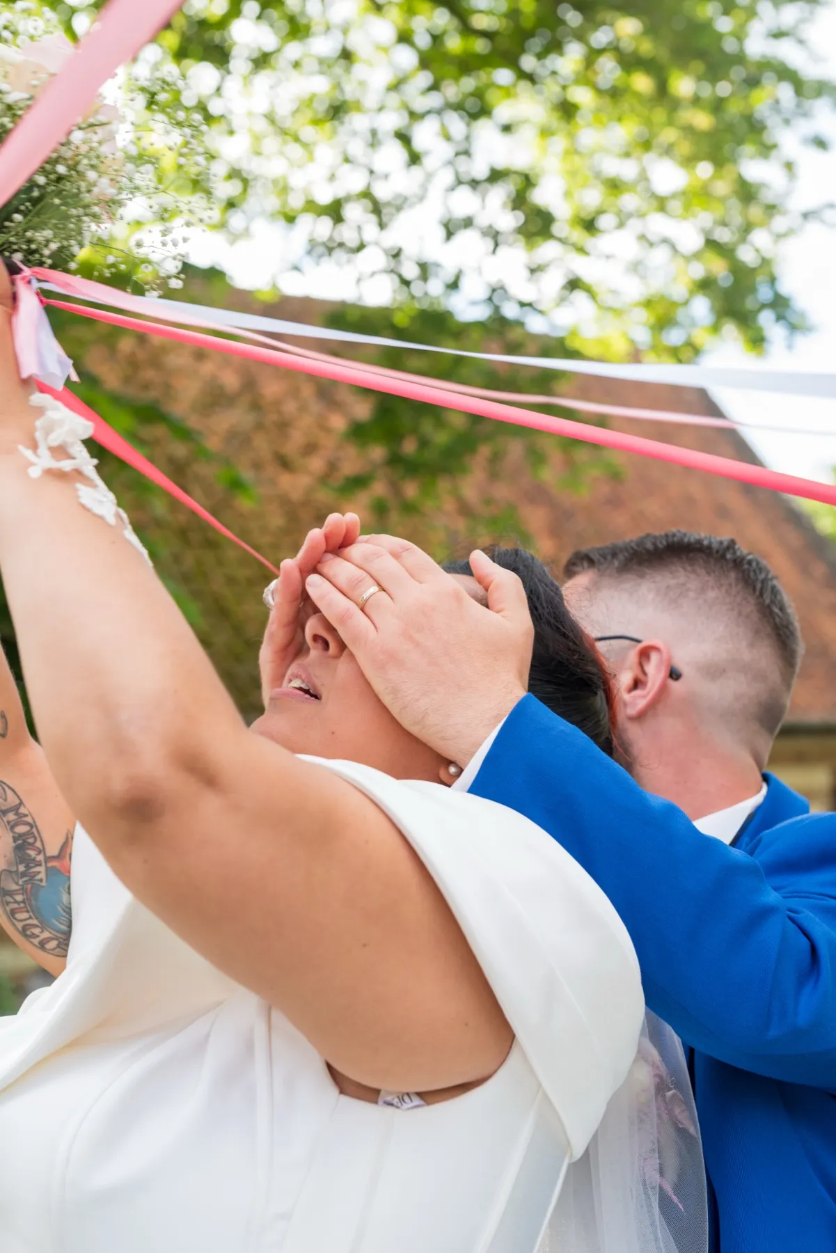 Le marié cache les yeux de la mariée qui tient le bouquet et ses rubans à bout de bras