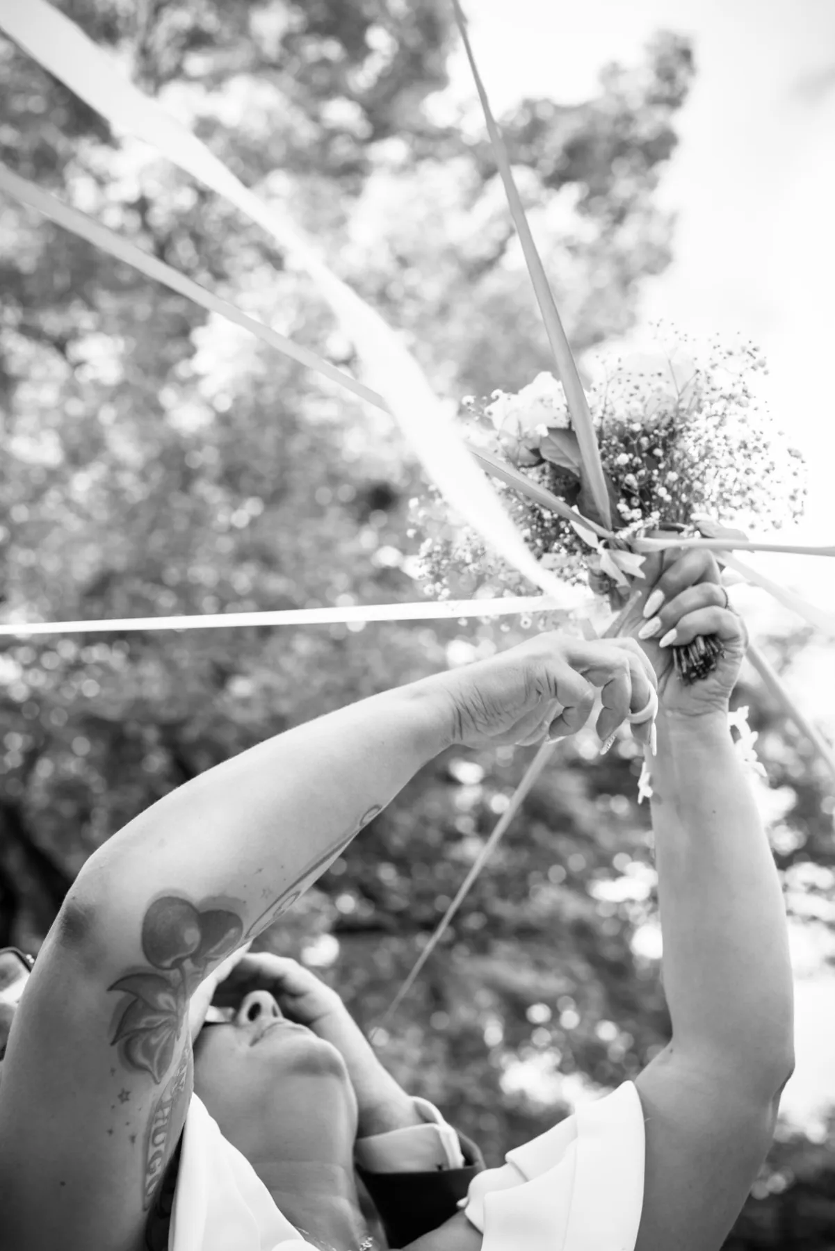 En noir et blanc, la mariée, les yeux cachés, lève le bouquet sous les rubans