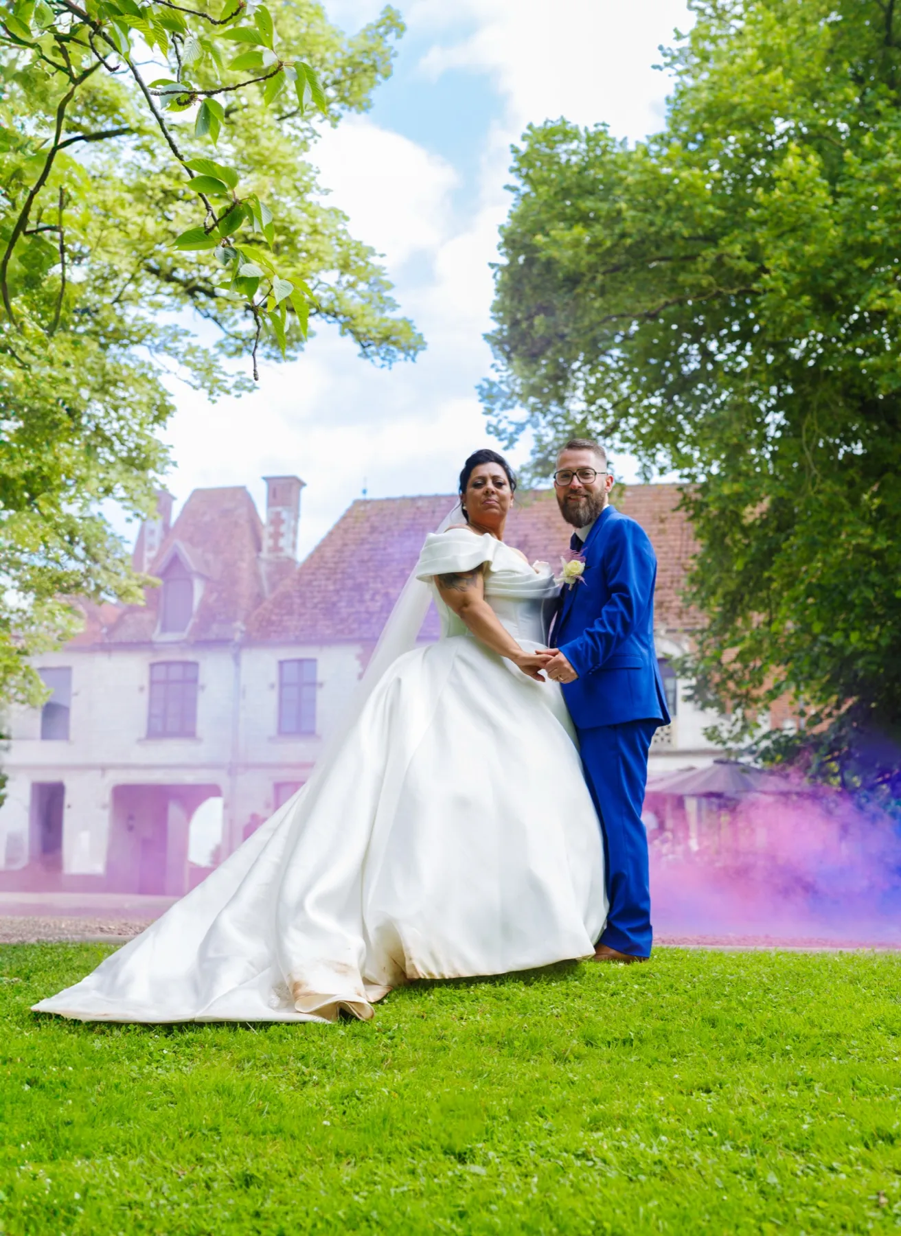 Les mariés se tiennent les mains sur une pelouse, dans un nuage de fumée violette, sous les arbres