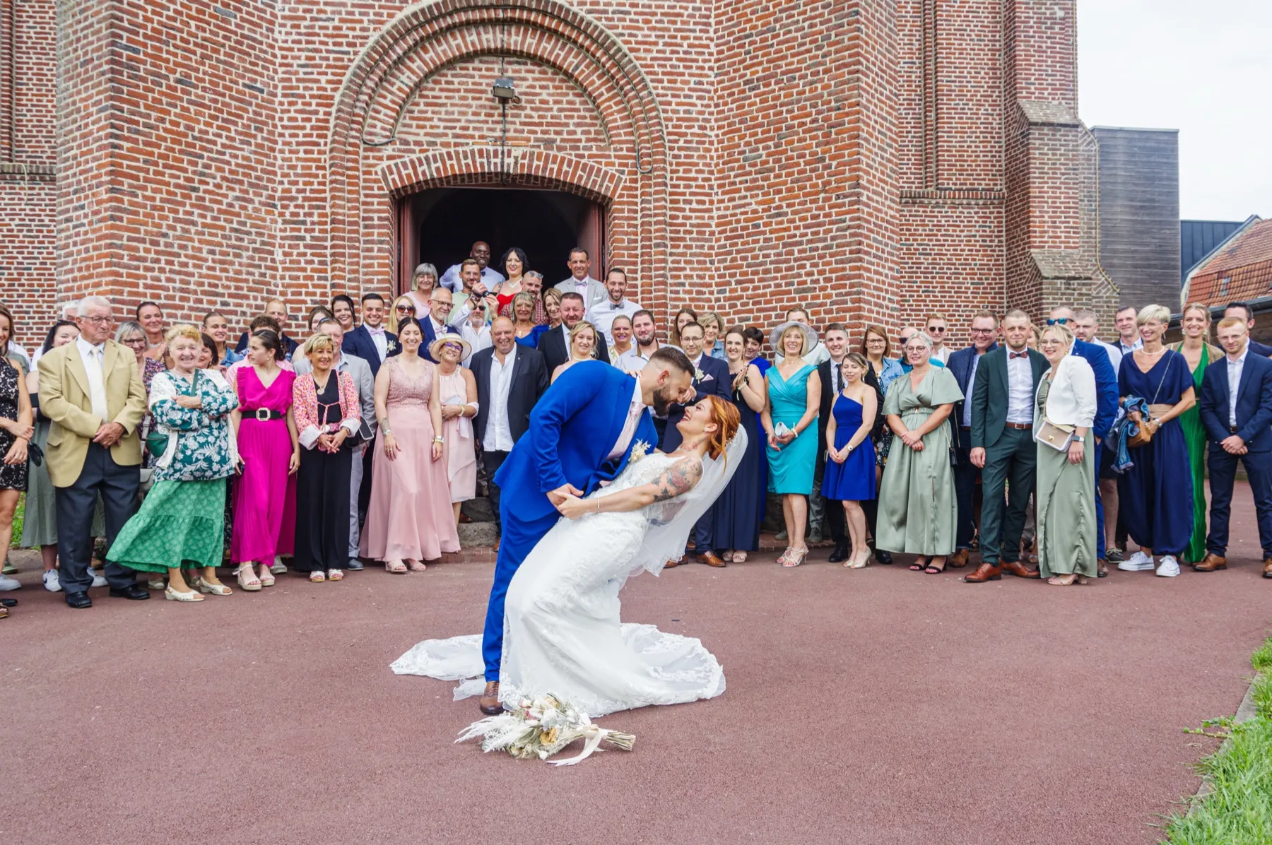 Devant l’église en briques, le marié renverse la mariée pour un baiser, entourés de tous leurs invités