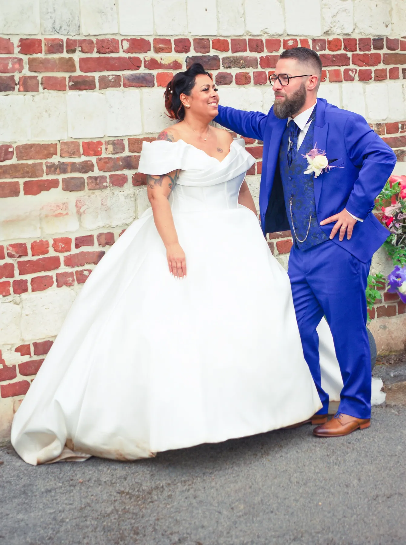 Contre un mur de briques, le marié pose la main sur l’épaule de la mariée qui rit