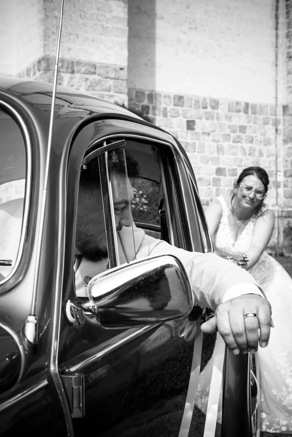 En noir et blanc, le marié à la fenêtre de la Coccinelle montre son alliance, la mariée rit derrière lui