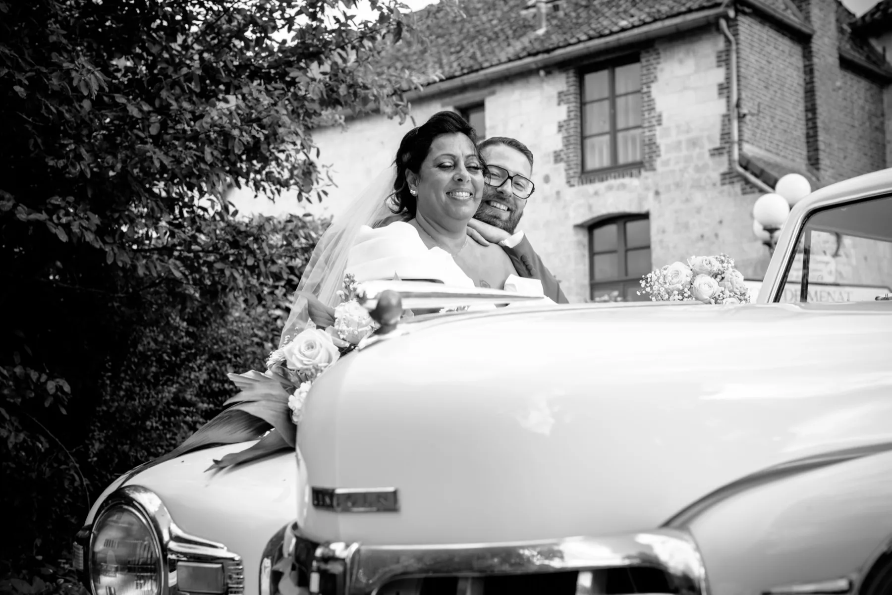 En noir et blanc, les mariés joue contre joue derrière le capot d’une voiture ancienne