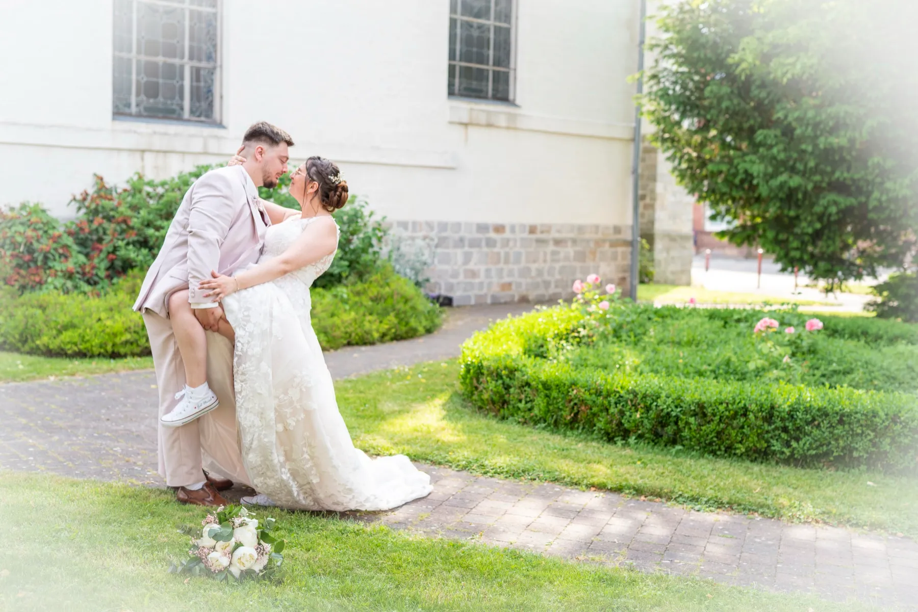 Sur une allée du jardin, le marié embrasse la mariée, le bouquet posé à leurs pieds
