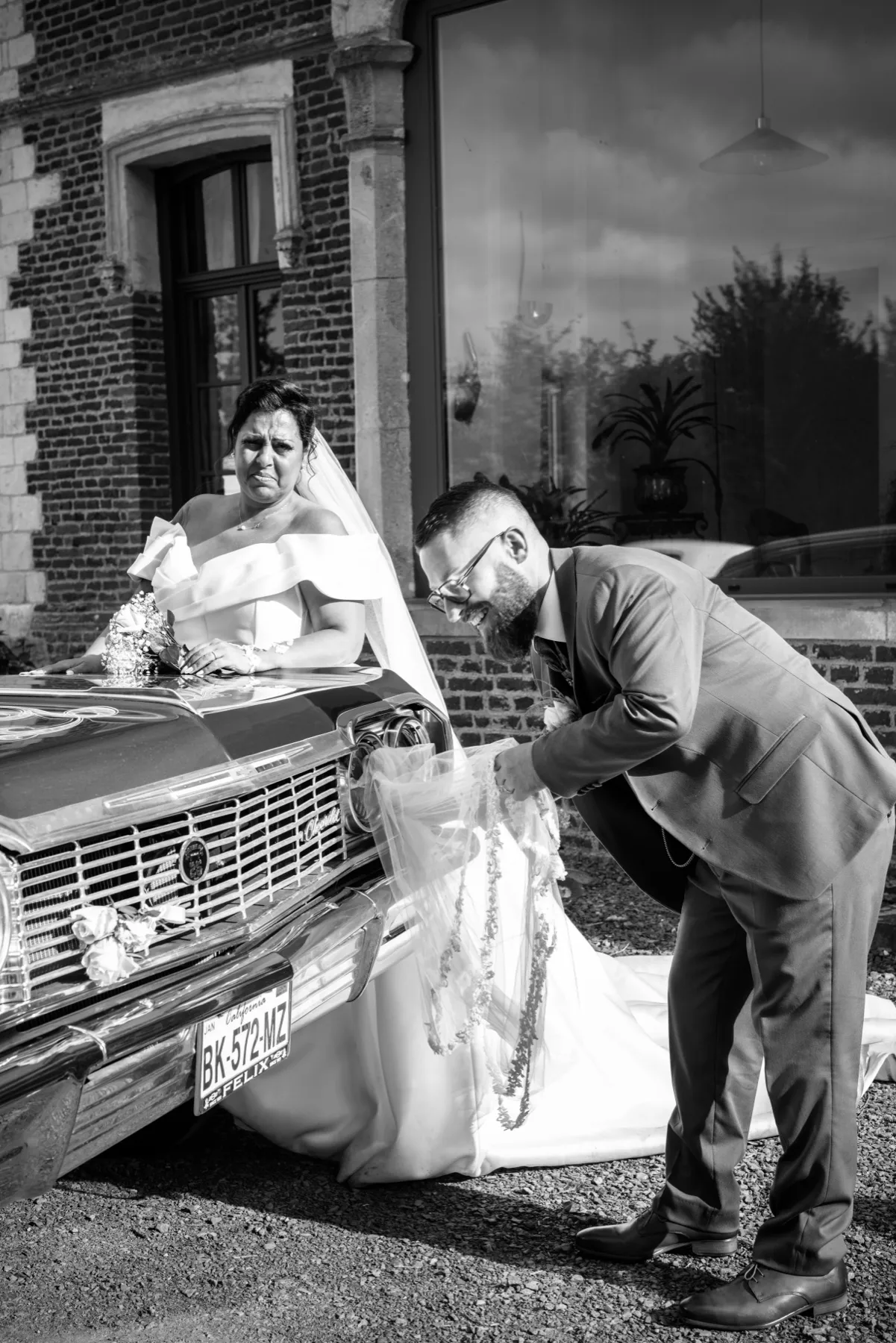En noir et blanc, le marié arrange la traîne de la mariée assise sur le capot d’une voiture ancienne