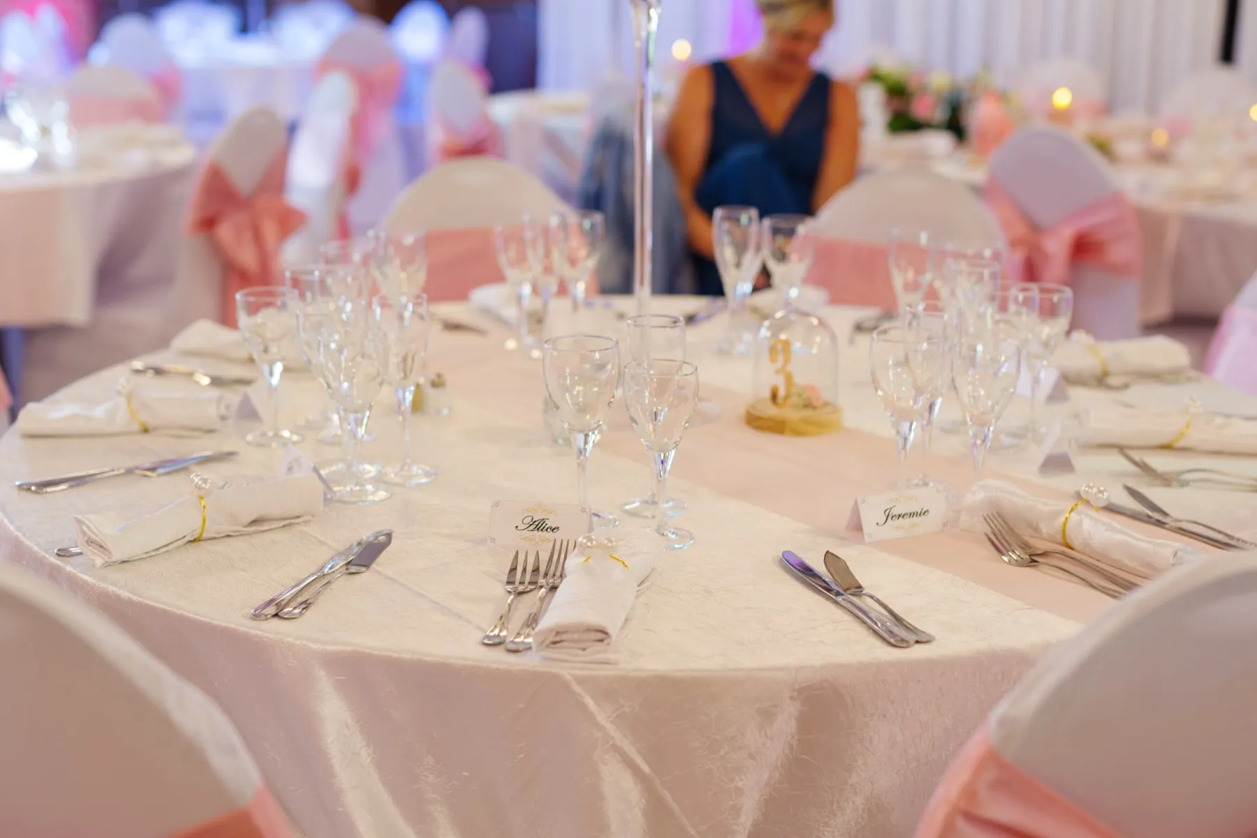 Une table ronde dressée, nappe satinée, verres en cristal et nœuds roses sur les chaises