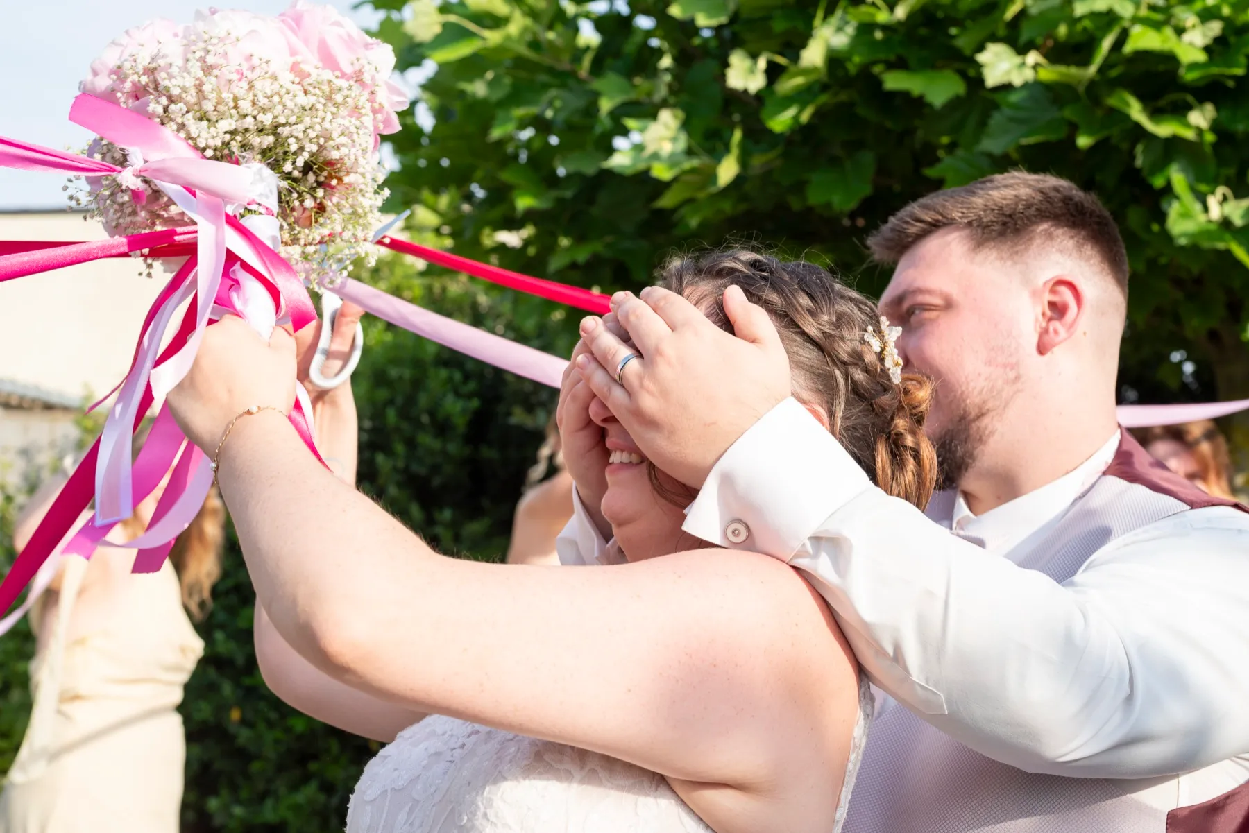 Le marié cache les yeux de la mariée qui tient le bouquet et ses rubans