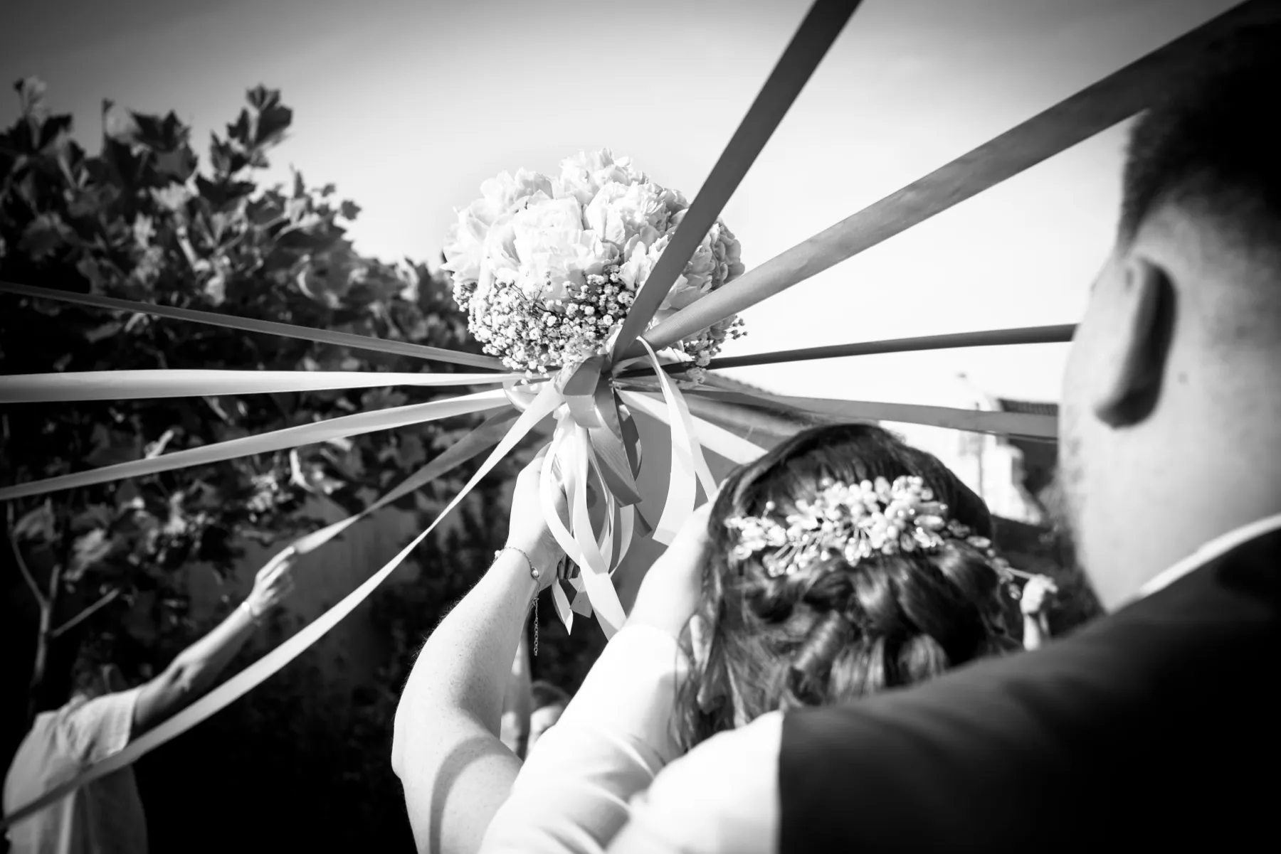 En noir et blanc, la mariée lève le bouquet sous les rubans tendus, le marié derrière elle