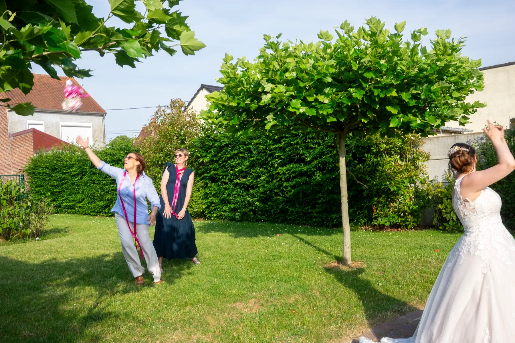 Dans le jardin, la mariée lève le bras vers deux invitées qui tiennent leurs rubans