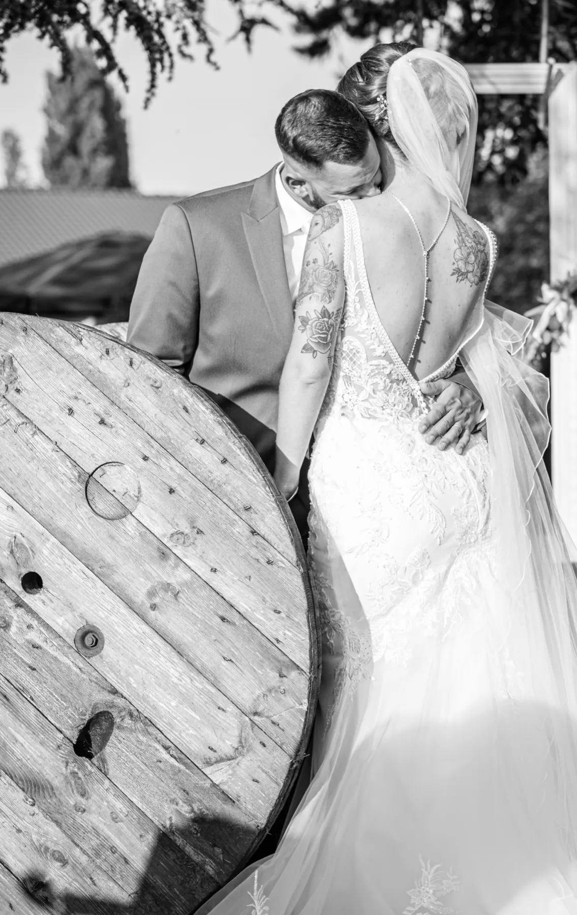 En noir et blanc, le marié embrasse l’épaule de la mariée, près d’un touret en bois