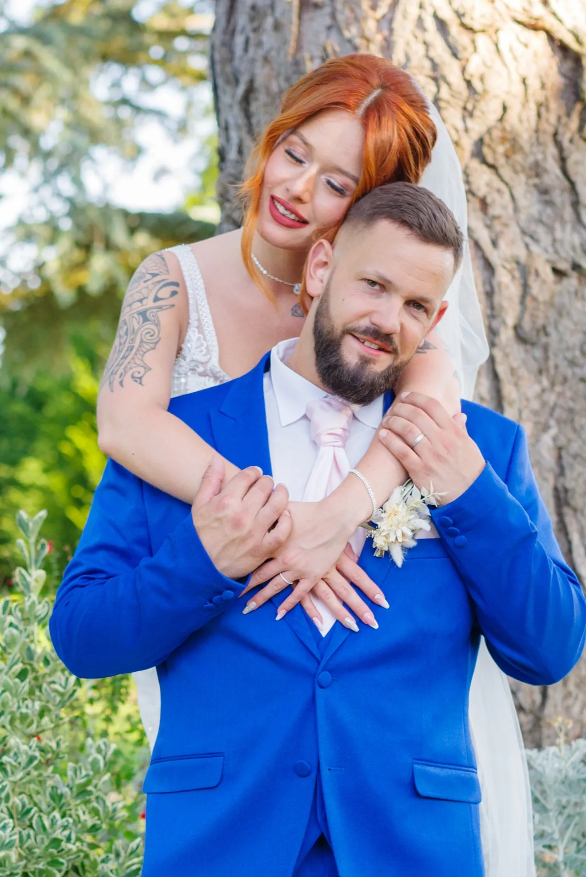 La mariée enlace le marié par-derrière, leurs mains jointes sur sa veste bleue