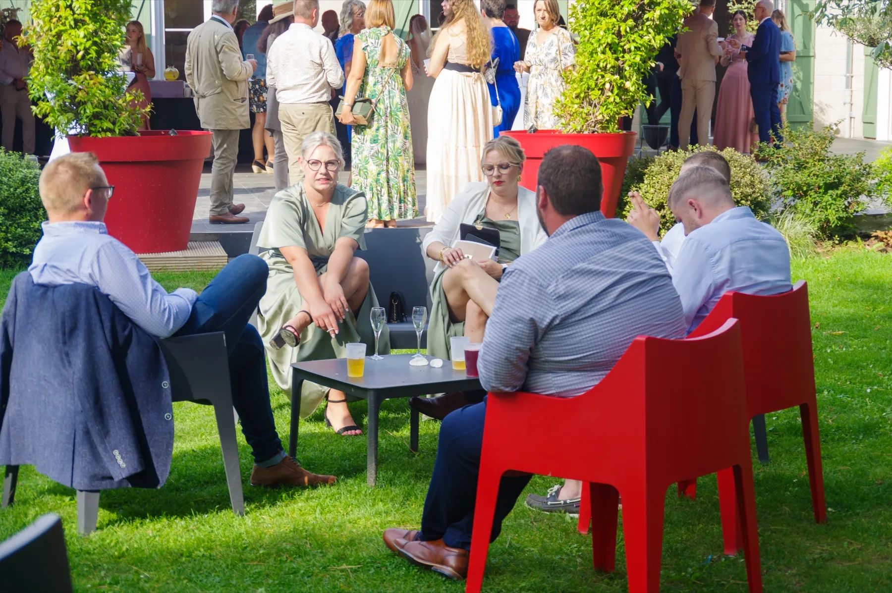 Des invités installés sur la pelouse autour d’un verre, dans des fauteuils rouges