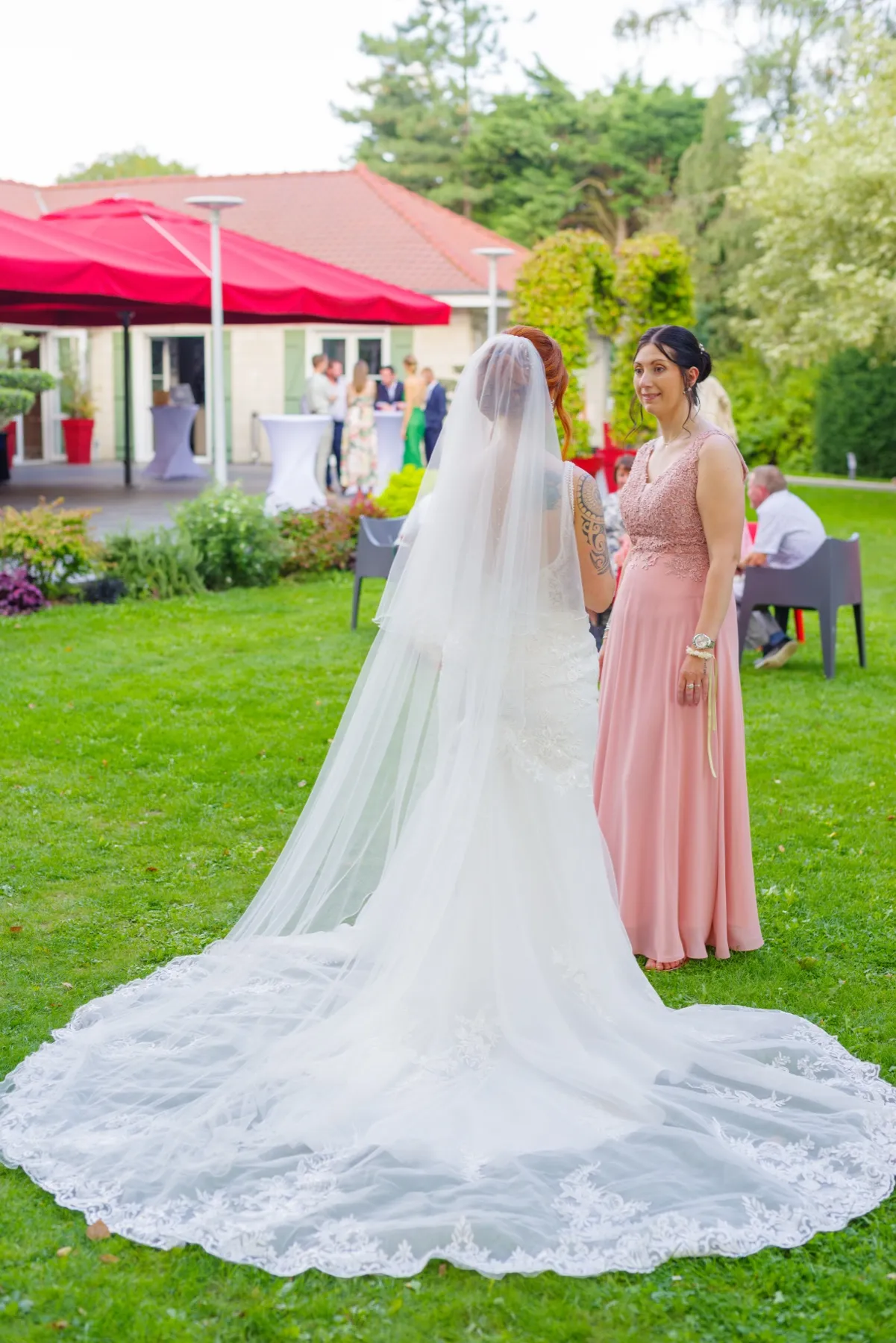 La mariée de dos, sa longue traîne de dentelle étalée sur l’herbe, parle avec une invitée