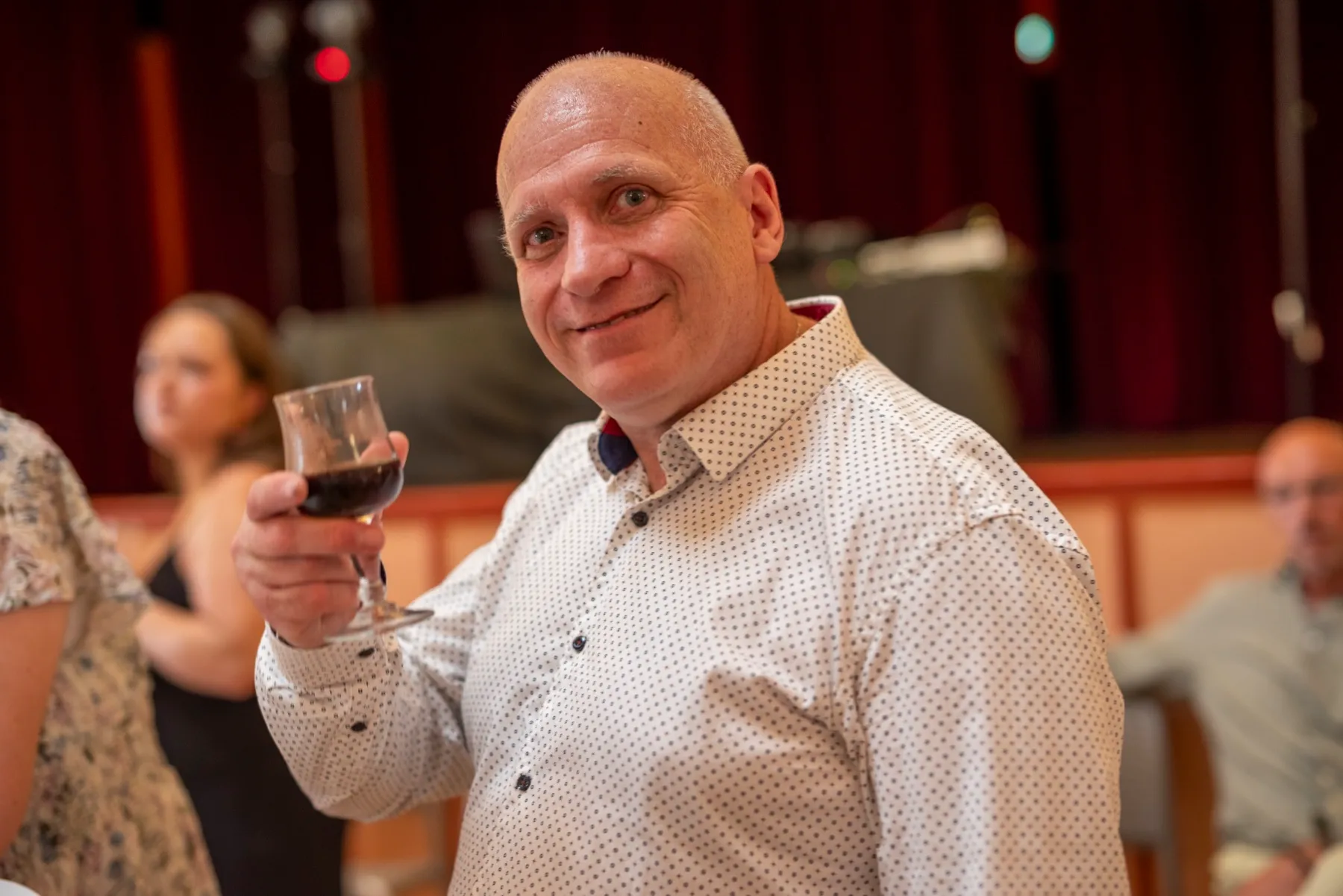 Un invité souriant lève son verre de vin rouge vers l’objectif