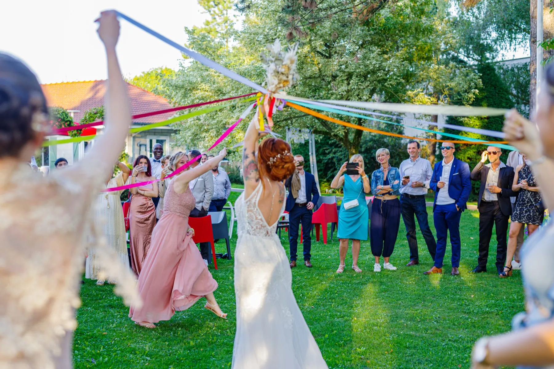 Vue de dos, la mariée tient le bouquet et ses rubans, les invités en cercle autour d’elle