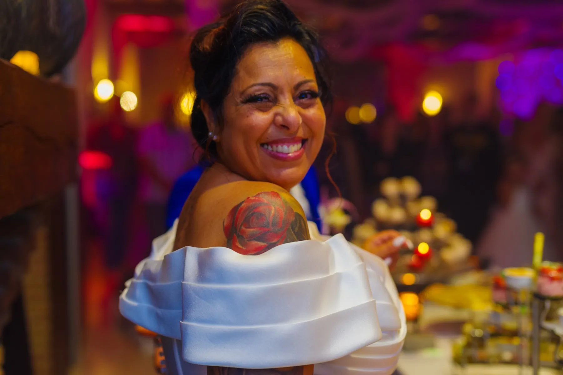 La mariée se retourne en souriant, une rose tatouée sur l’épaule, devant le buffet illuminé