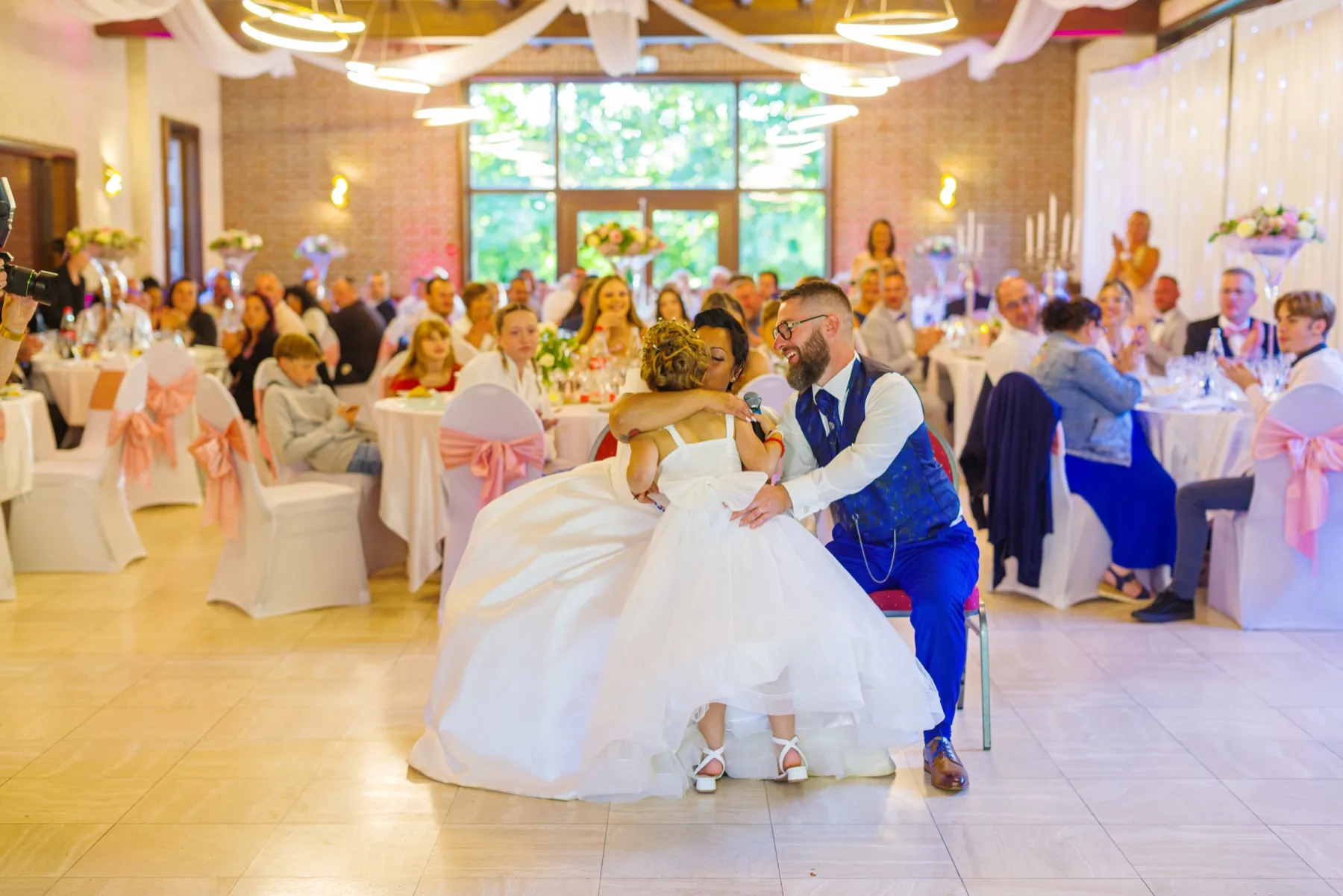 La mariée, assise sur les genoux du marié, l’enlace, un micro à la main, devant les invités attablés