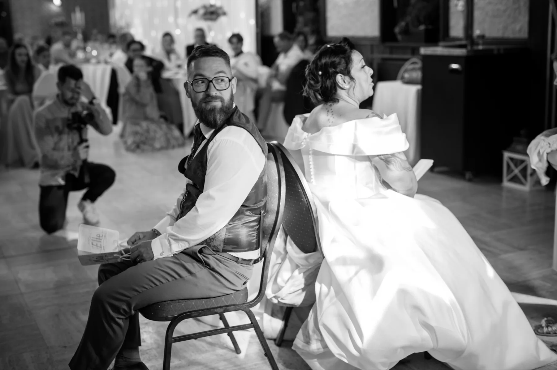 En noir et blanc, les mariés assis dos à dos sur deux chaises, un invité accroupi les photographie