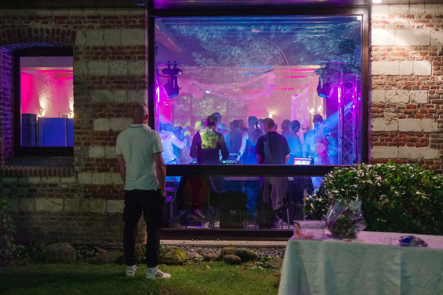 De nuit, vu du jardin, un homme regarde la piste de danse éclairée par la fenêtre